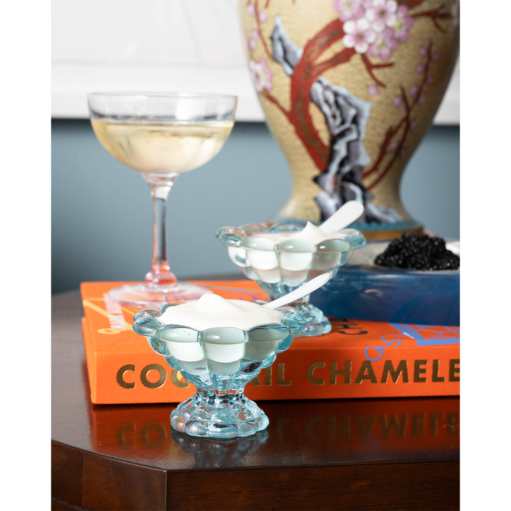 White shell-shaped glass spreader resting on clear footed glass dish on wooden table with books and cocktail glass in.