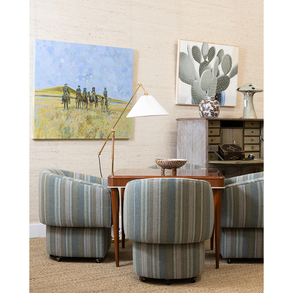 A mid-century modern living room setup featuring four striped armchairs in muted blue, green, and beige tones around a wooden table. The room is styled with a floor lamp, a sideboard cabinet, and two framed wall art prints, including a cactus painting.
