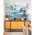 A large abstract canvas painting in shades of blue, white, and beige hangs above a mid-century modern sideboard with yellow wood cabinets. The room is styled with blue and white ceramic vases, potted pink and white flowers, and a light blue table lamp on top.