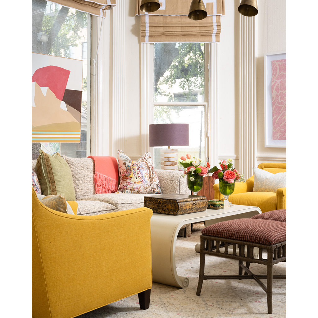 A styled living room interior featuring a mustard yellow armchair, a cream sofa with patterned and coral pillows, a curved cream coffee table, a brown upholstered stool, and floral arrangements by tall windows with beige blinds.