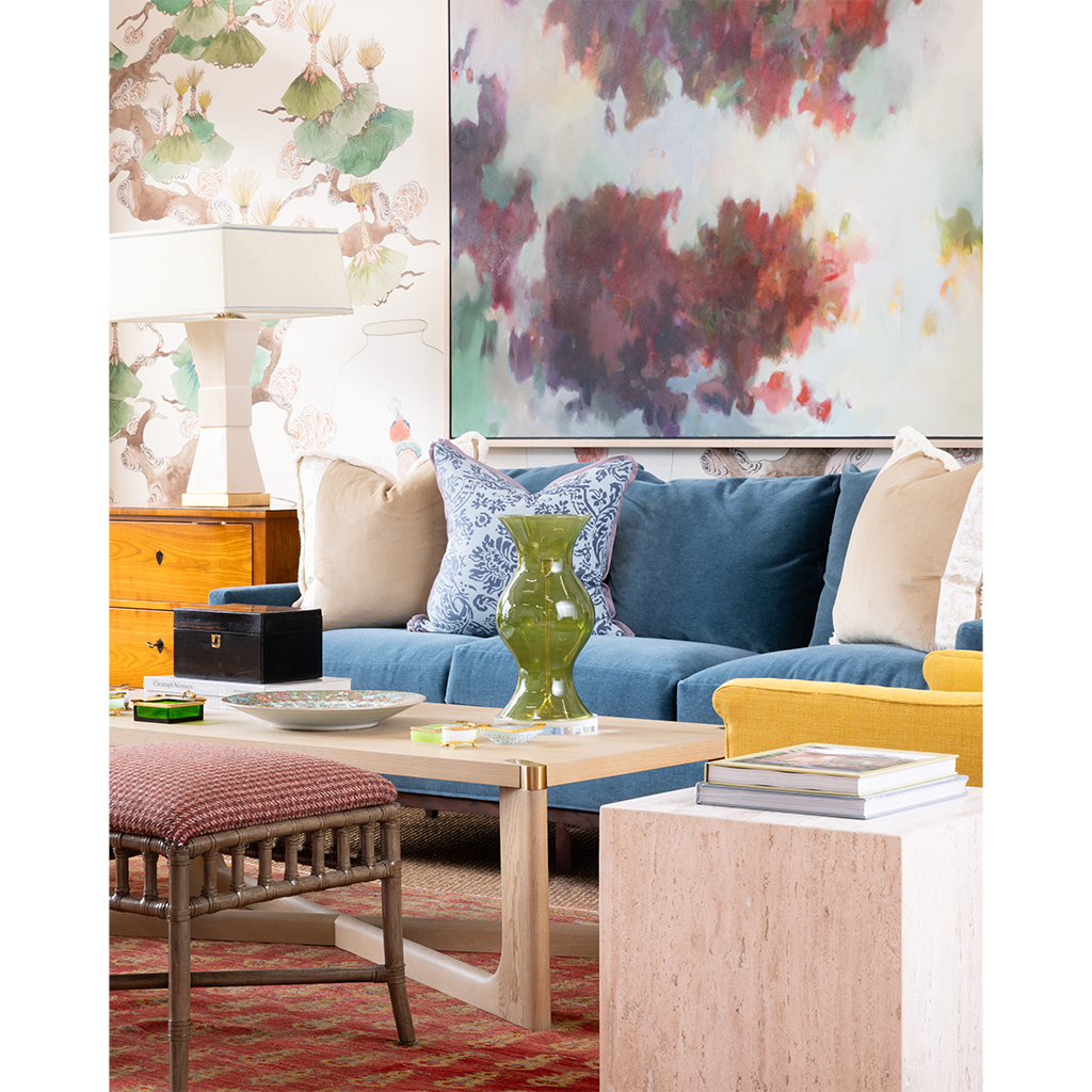 A styled living room scene featuring a blue sofa with beige and blue patterned pillows, a yellow accent chair, a light wood coffee table, and a large abstract painting on the wall behind it.