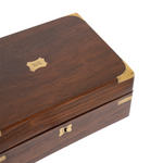 A dark brown wooden box with brass corner protectors and a small brass crest on the lid, shown on a white background.