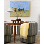 A mid-century modern living room scene featuring a large square canvas painting of soldiers on horseback in a field, hung on a white textured wall above a wooden console table. The room is furnished with striped armchairs in muted green, blue, and beige tones, a yellow throw blanket, and a brass floor lamp.