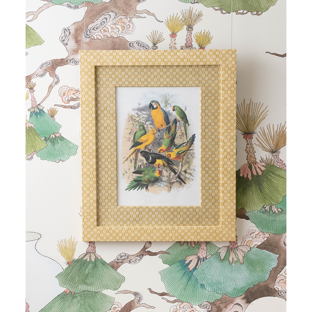 A framed bird illustration print with a patterned gold and beige frame, displayed against a matching botanical wallpaper featuring green leaves and brown branches.