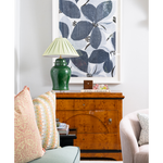 A styled interior scene featuring a green ceramic table lamp on a wooden sideboard, with patterned pillows, a light-colored armchair, and a large framed floral artwork on a white wall.