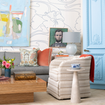 An elevated, eclectic living room vignette featuring a cream textured-stripe upholstered swivel chair with a coral throw over one arm, beside a round travertine pedestal side table holding a blue-and-white teacup. A gray sofa layered with an embroidered pagoda-motif pillow and coral velvet bolster sits behind a zebrawood coffee table topped with an antique lacquered box and a vase of pink and yellow flowers, all set against floral line-drawing wallpaper, framed artwork, and a powder-blue paneled door.