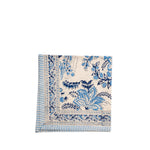 A folded cream-colored cotton napkin with a blue floral and botanical print, featuring a striped border on the left side, set against a plain white background.