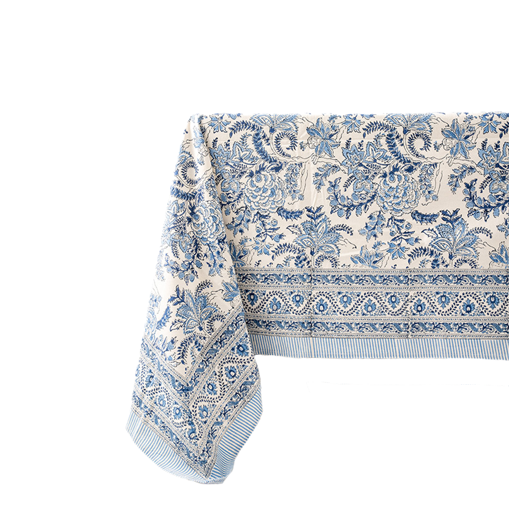 A cream-colored tablecloth with an intricate blue floral and botanical pattern, featuring a decorative border and striped fringe, shown draped on a plain white background.