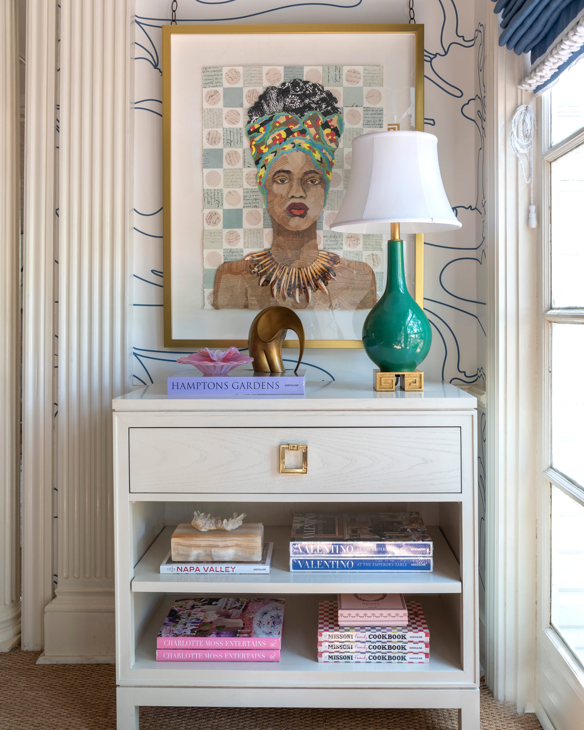 Interior with side table filled with books, a green and white lamp, and a Brenda Bogart collage