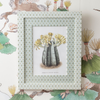 A framed botanical art print featuring a painted illustration of three gray pubeck trees with yellow flowers, set in a patterned white frame against a matching illustrated wallpaper background.