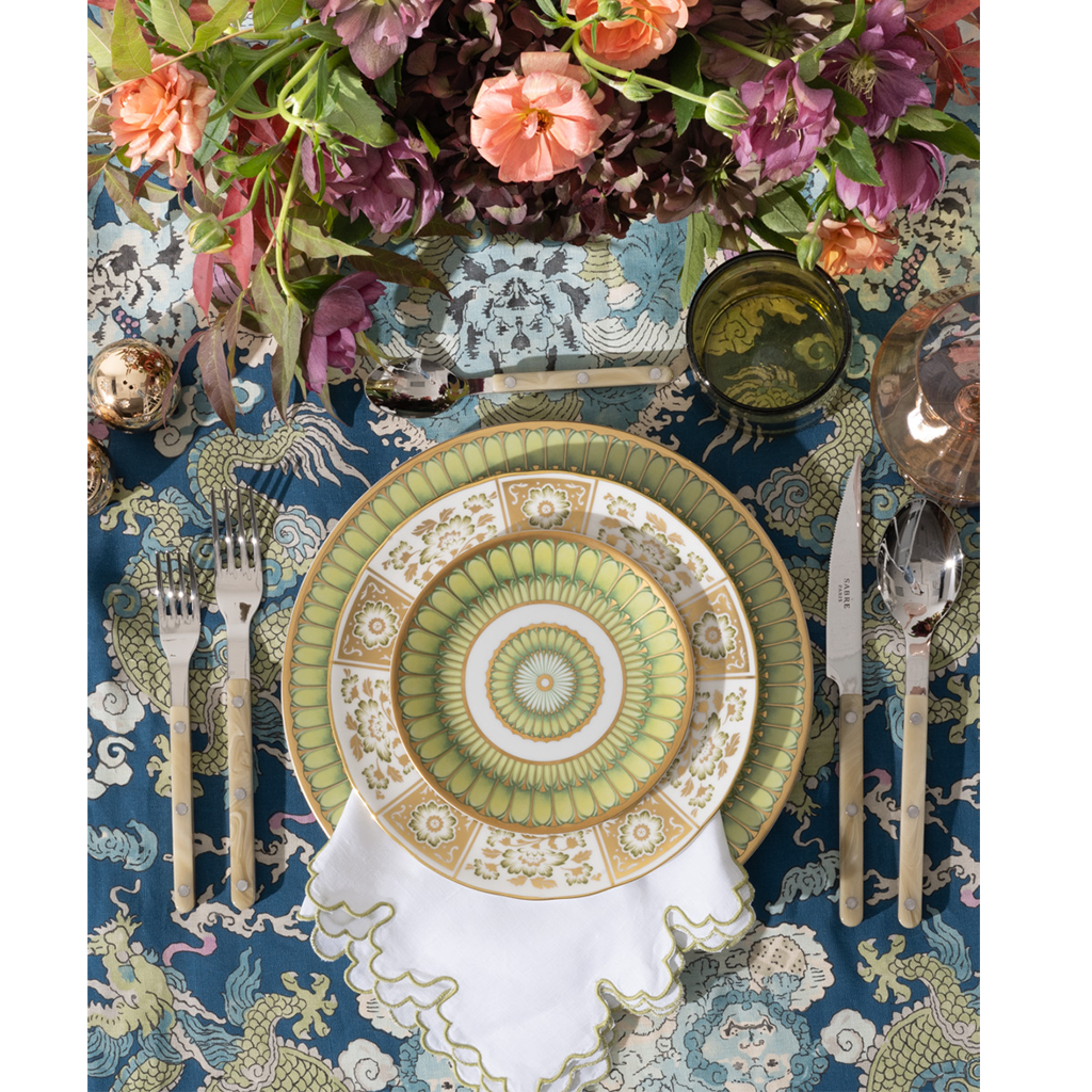Blue tablecloth with intricate white botanical and animal patterns under floral centerpiece and patterned dinnerware set.