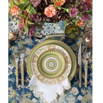 Blue tablecloth with intricate white botanical and animal patterns under floral centerpiece and patterned dinnerware set.