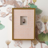 A square framed wall art piece with a gold-toned frame, pale pink patterned mat, and a small central vase sculpture, displayed against a decorative botanical wallpaper.