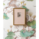 A framed wall art piece with a gold frame and a pink patterned backing, mounted on a white wallpapered wall with botanical illustrations.