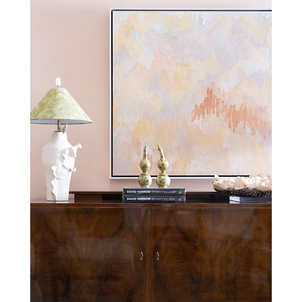 A framed abstract wall art print hangs above a dark wood console table in a styled interior. The artwork features soft pastel washes of beige, cream, and peach with a coral-like textured accent.
