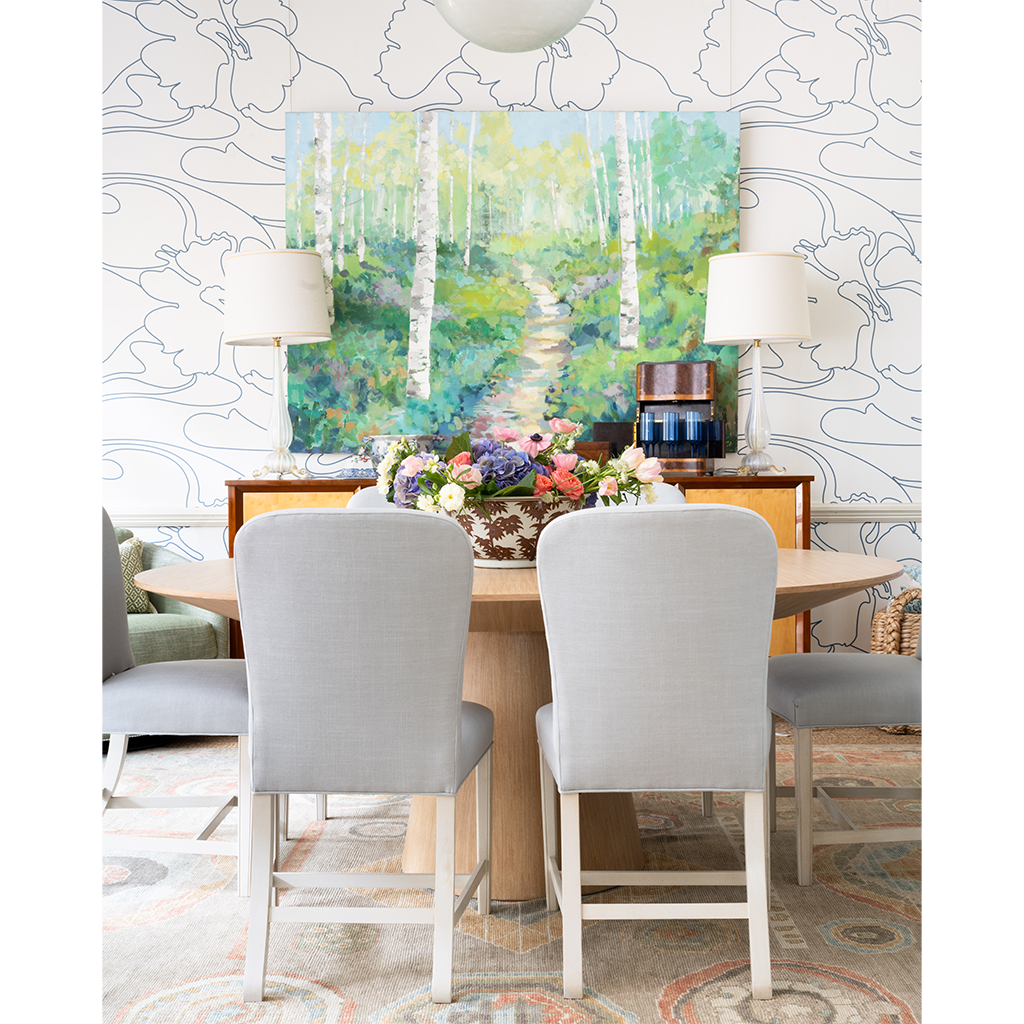 A styled dining room scene with a round wooden table, four light gray upholstered chairs, two table lamps, and a large colorful forest painting mounted on the wall behind. The room has patterned wallpaper, a rug, and a sideboard with decorative objects.