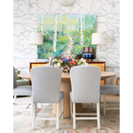 A styled dining room scene with a round wooden table, four light gray upholstered chairs, two table lamps, and a large colorful forest painting mounted on the wall behind. The room has patterned wallpaper, a rug, and a sideboard with decorative objects.
