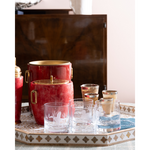 A bar-themed product display featuring a red and gold ice bucket set and matching clear glass tumblers with gold trim, arranged on a decorative tray in an indoor setting.