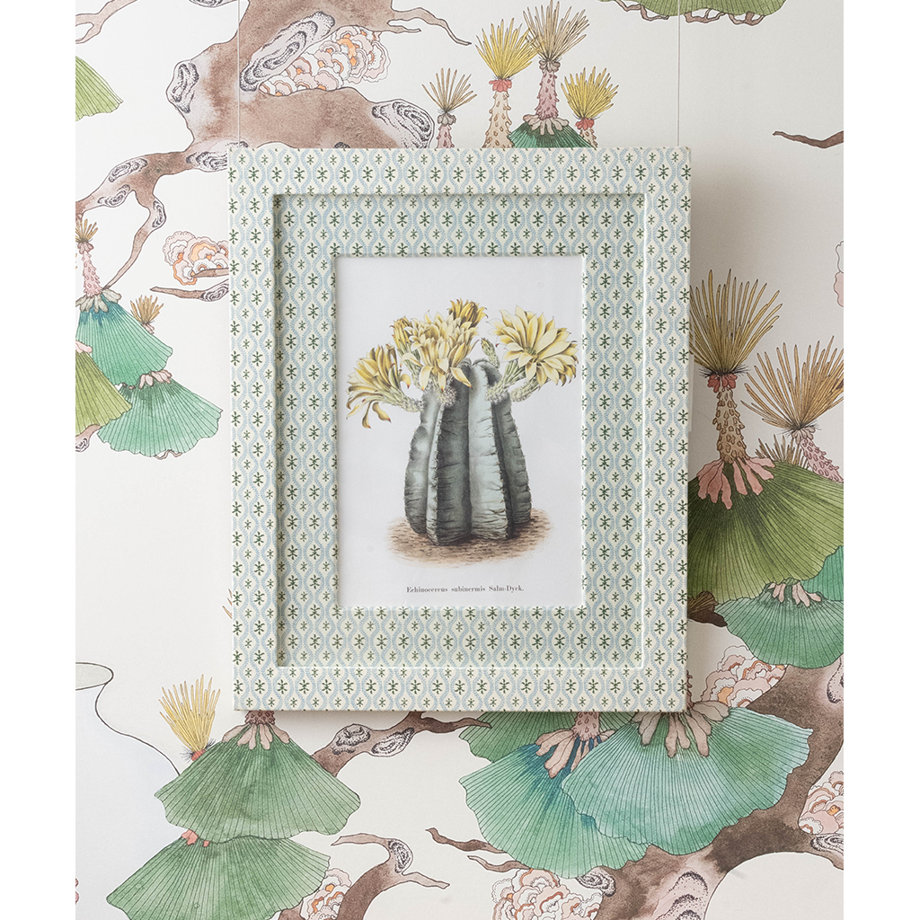 A framed botanical art print featuring a striped cactus with yellow flowers, set against a patterned wallpaper with matching green and beige illustrations.