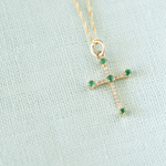 Gold cross necklace with green gemstones on a light blue fabric background