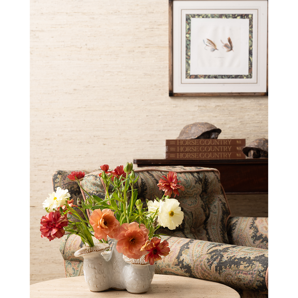 A styled interior scene with a floral arrangement in a ceramic vase on a table in the foreground, an upholstered armchair behind it, books, and framed wall art on a textured beige wall.