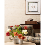 A styled interior scene with a floral arrangement in a ceramic vase on a table in the foreground, an upholstered armchair behind it, books, and framed wall art on a textured beige wall.