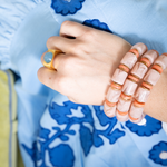 A close-up of a person's hand and wrist wearing a stack of peach and white marbled beaded bracelets with gold-toned spacer rings, along with a gold ring with a blue stone, set against a light blue embroidered garment.
