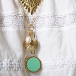 A gold-tone layered necklace with a textured sunburst pendant, a white pearl, and a mint green oval pendant, displayed on a white lace-trimmed fabric.