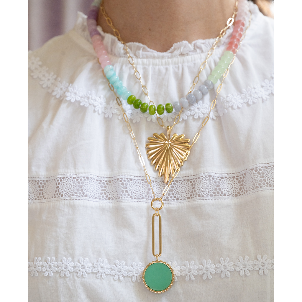A gold-tone layered necklace with a textured sunburst pendant, a white pearl, and a mint green oval pendant, displayed on a white lace-trimmed fabric.