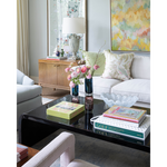 Glossy black coffee table with stacked books and pink flowers in vases in a bright living room setting.