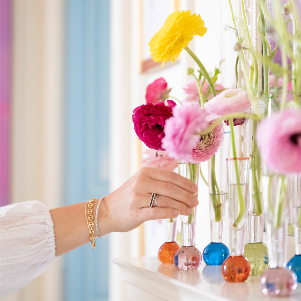 Blue glass flute vase held by hand with gold bracelet, filled with colorful flowers.
