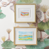Two small framed abstract watercolor paintings hang on a white wallpaper with a botanical pattern. The frames are light wood with white matting.