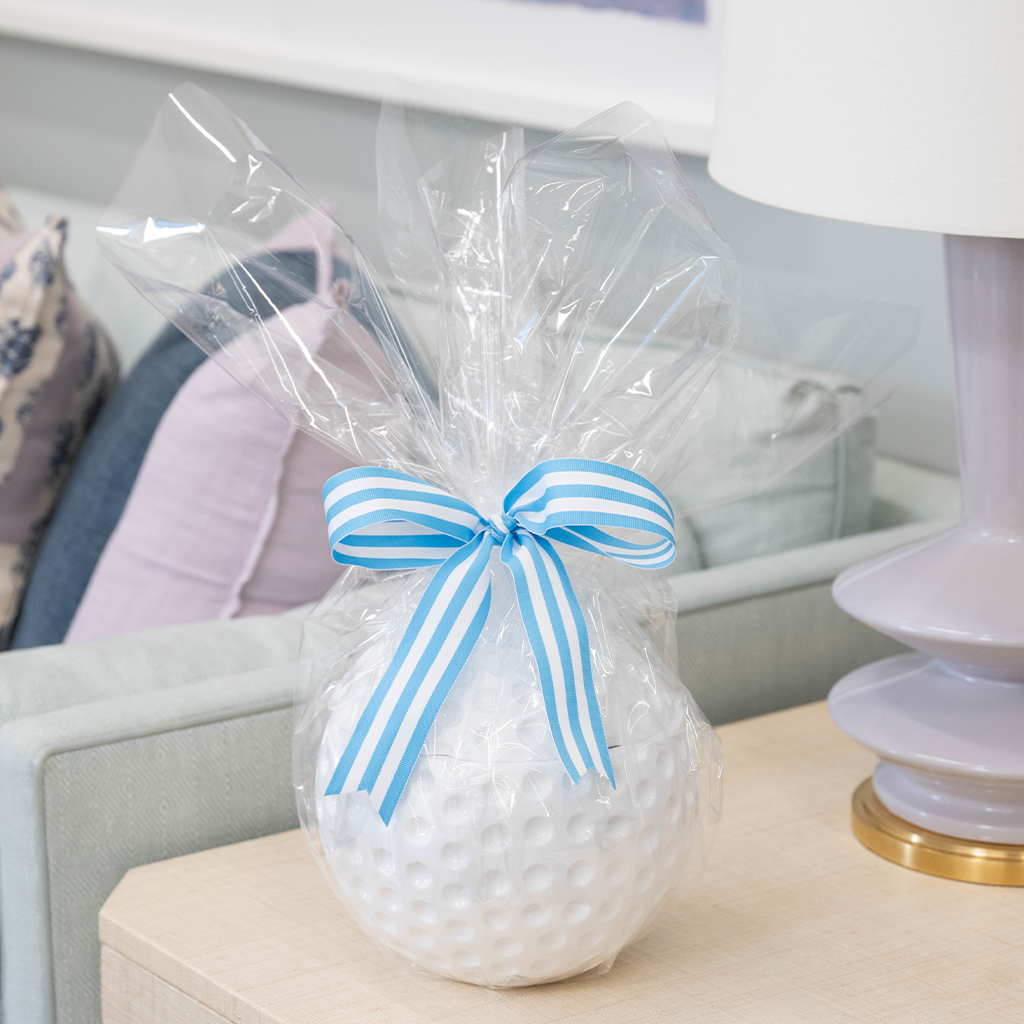 White golf ball-shaped ice bucket wrapped in clear plastic with blue and white striped bow on wooden table.