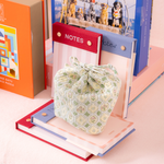 A small drawstring pouch with a sage green and cream geometric pattern sits on top of a stack of pastel-colored books, with stationery items like a red 'NOTES' pad and a blue notebook in the background.
