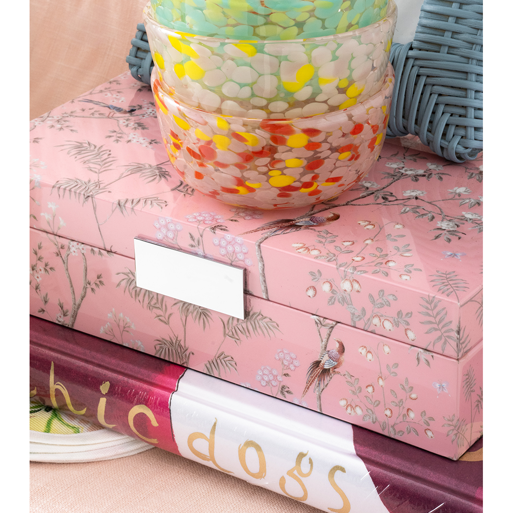 Stack of colorful books with a decorative pink box featuring floral patterns on a beige surface.