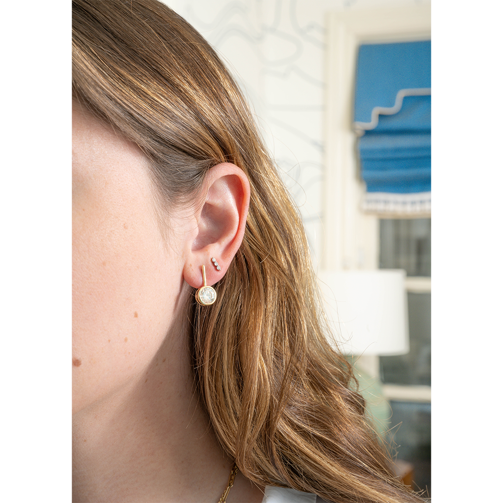 A close-up side profile of a woman wearing a delicate gold hoop earring with a small round white pearl at the front, along with small black and silver studs in her ear cartilage. Her light brown hair is visible, and the background shows a softly blurred interior room with a blue window shade.