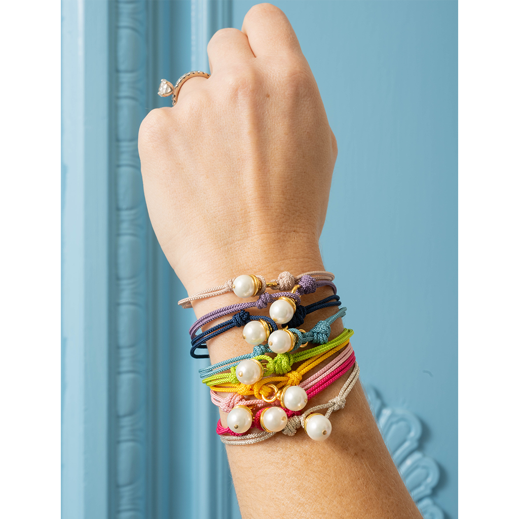 A close-up of a person's wrist wearing a stack of colorful cord bracelets with white pearl beads, set against a light blue paneled background.