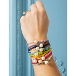 A close-up of a person's wrist wearing a stack of colorful cord bracelets with white pearl beads, set against a light blue paneled background.