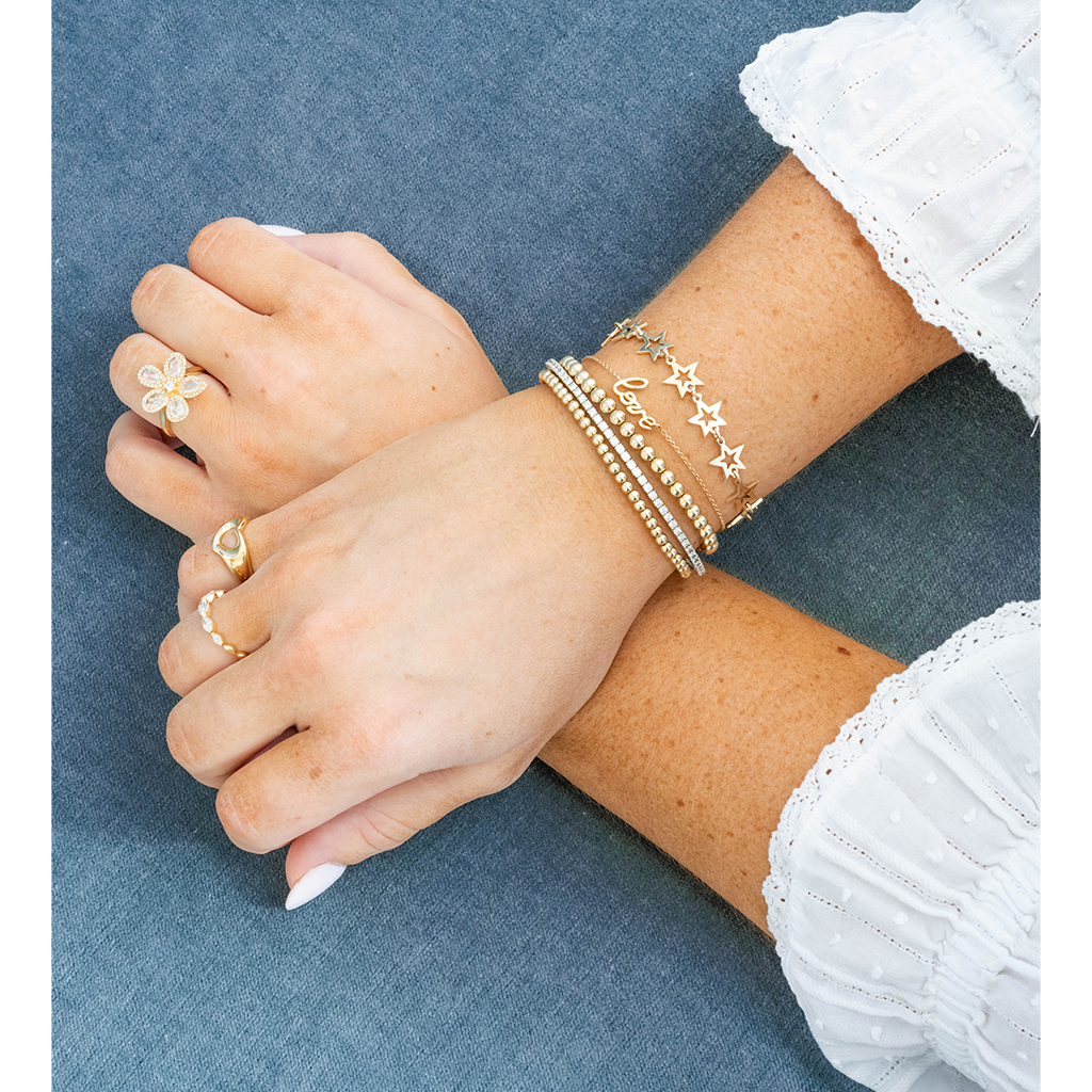 A close-up of crossed hands and forearms wearing delicate gold jewelry, including a flower-shaped ring, a small band ring, and several stacked bracelets, against a textured blue fabric background.