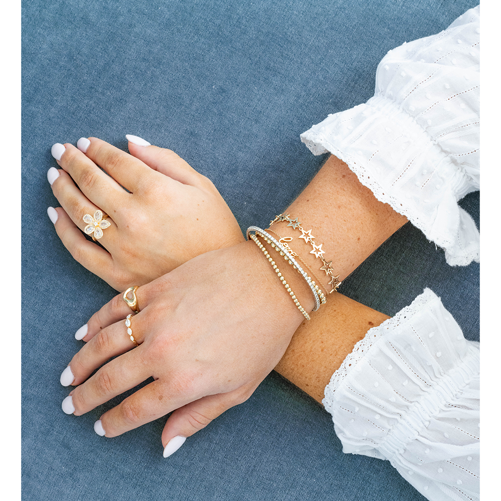 A close-up of crossed hands and forearms wearing delicate gold jewelry, including a flower-shaped ring, a small band ring, and several stacked bracelets, against a textured blue fabric background.