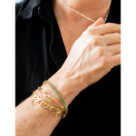 A close-up of a person's wrist wearing multiple gold bracelets, including a nameplate bracelet, a green stone bracelet, and a slim gold chain, with a matching delicate necklace also visible.