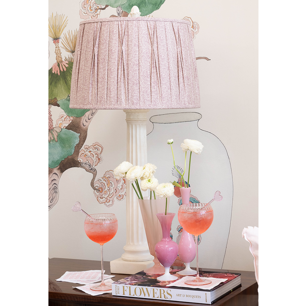 White ceramic table lamp with pleated pale pink fabric shade on floral-themed background.