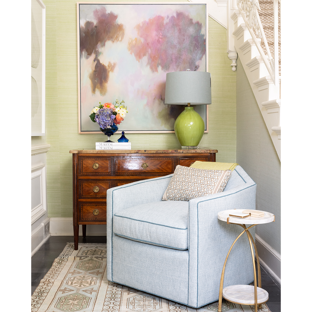 A light blue upholstered armchair in a styled living room corner, next to a small marble-top side table and above a wooden console table with a green ceramic lamp and framed abstract wall art.