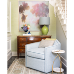A light blue upholstered armchair in a styled living room corner, next to a small marble-top side table and above a wooden console table with a green ceramic lamp and framed abstract wall art.