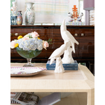 A pair of white ceramic bird sculptures displayed on stacked blue books on a light wood table, with a glass vase of flowers and decorative vases in the background.