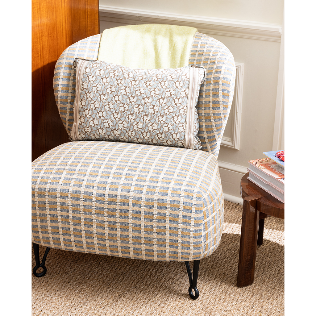 A classic wingback armchair upholstered in a beige, blue, and white plaid fabric, styled with a matching patterned lumbar pillow and a pale yellow throw blanket. The chair is set in a cozy room next to a small wooden side table.
