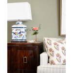 Decorative lamp on a wooden side table with a vase of flowers and a floral-patterned pillow on a sofa.