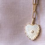 Heart-shaped pendant with a diamond on a gold chain against a textured beige background
