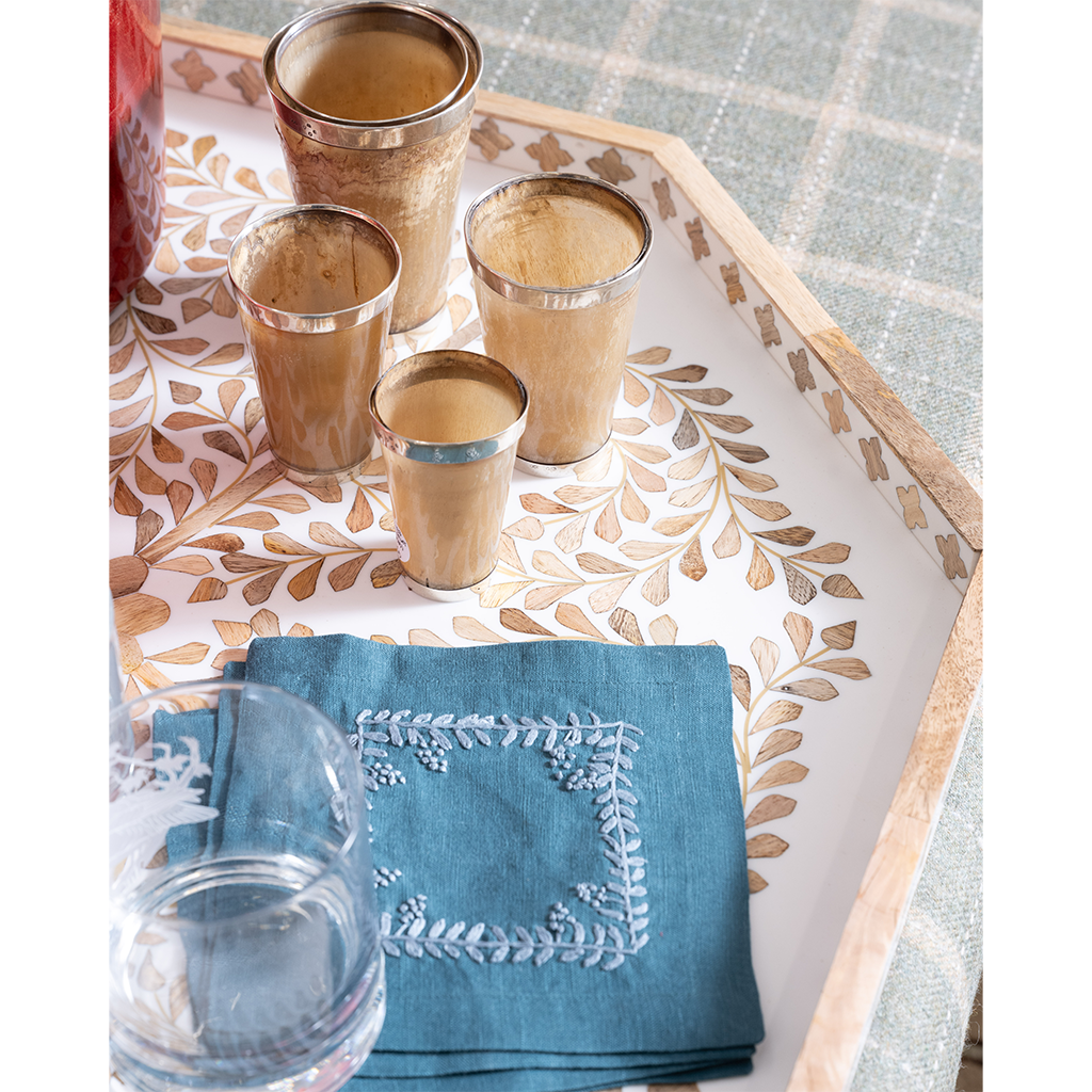 A styled tabletop scene with a decorative rectangular tray holding four metallic gold shot glasses, a folded blue napkin with white embroidered border, and part of a clear drinking glass. The tray has a leaf pattern and sits on a blue plaid tablecloth.