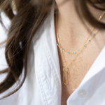 Delicate gold keyhole chain necklace layered with a thin blue gemstone chain on a woman wearing white shirt.